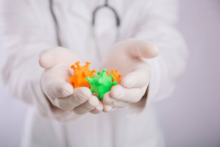 A doctor in a white coat and gloves on a white background holding a mock coronavirus in his hands....