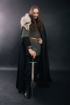 Warrior in armor with a sword. A guy with long hair and a beard, a fox collar. Dressed in chain mail, a black cloak and black pants holds a sword in his hands.の写真素材
