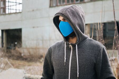 A guy in a gray sweater with a blue medical mask. Holds a hood with his own hands. Against the background of destroyed and abandoned buildings. Quarantined man.の写真素材