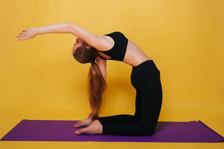 Athletic, beautiful young girl practicing yoga doing ushtrasana pose. Wear black sportswear on a plain yellow background.の写真素材