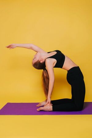 Athletic, beautiful young girl practicing yoga doing ushtrasana pose. Wear black sportswear on a plain yellow background.の写真素材