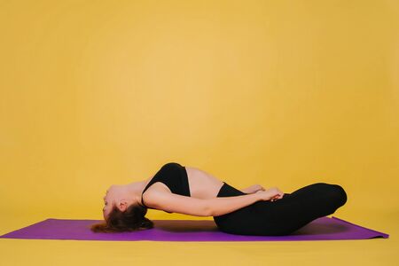 Athletic, beautiful young girl practicing yoga, doing Padma Matsiasana pose. Wear black sportswear on a plain yellow background while standingの写真素材