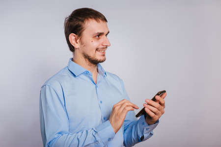 A guy in a blue shirt holds a smartphone on a white background. A male office worker is typing text on the phone.の写真素材