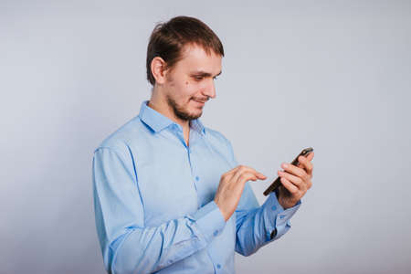 A guy in a blue shirt holds a smartphone on a white background. A male office worker is typing text on the phone.の写真素材