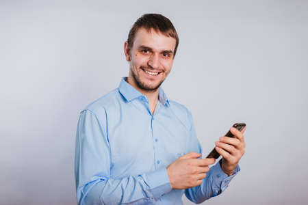 A guy in a blue shirt holds a smartphone on a white background. A male office worker is typing text on the phone.の写真素材
