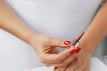 Woman hands - nail polish on light background. Girl holding nail polish brush in her hand. She painting her nails in red colourの写真素材
