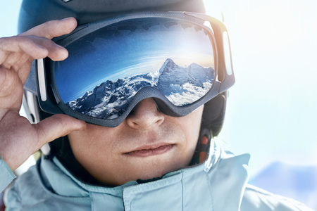 Close up of the ski goggles of a man with the reflection of snowed mountains.  A mountain range reflected in the ski mask.  Portrait of man at the ski resort on the background of mountains and skyの写真素材