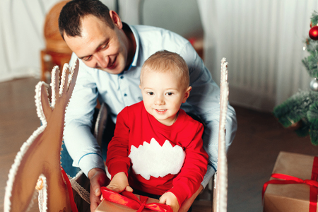 Happy father with his cute and smiling baby sitting on the chair near Christmas tree. New Year Celebration. Decorations.  Xmas eventの写真素材