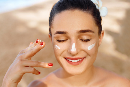 Beautiful Woman smile applying sun cream on face. Skin and  Body  care. Sun protection. Girl in bikini holding sunscreen bottle on the beach. Female in hat smear moisturizing lotion on skinの写真素材