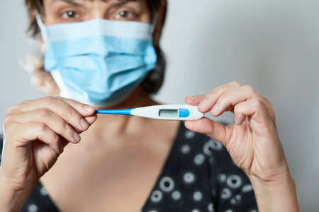 Woman in a mask with Coronavirus reads his temperature on a thermometer. Female measures the temperature. Check body temperature for virus symptoms. Copy space. Coronavirus. 2019 Novel Coronavirus (2020-nCoV)の写真素材