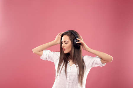 Beautiful woman smiling looking aside with smartphone in hands enjoying favorite music, via wireless headphones over pink background. Girl uses wireless earphones and dancing ..の写真素材