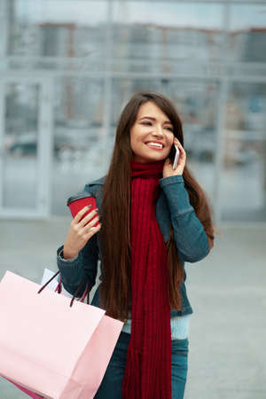 Young woman with shopping bags walking out from shop and talking on the phone. Woman with coffee. Consumerism, shoppingの写真素材