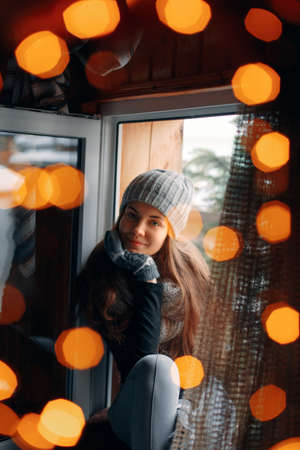 Beautiful woman sitting in a sweater home by the window. Blurred winter snow tree background. Morning, coziness, winter and people concept. Bright lightsの写真素材