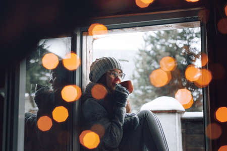 Beautiful woman holding and drinking a cup of coffee or cocoa in gloves sitting home by the window. Blurred winter snow tree background. Morning, coziness, winter and people conceptの写真素材