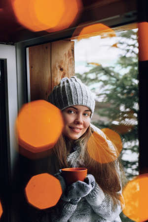 Beautiful woman holding and drinking a cup of coffee or cocoa in gloves sitting home by the window. Blurred winter snow tree background. Morning, coziness, winter and people conceptの写真素材