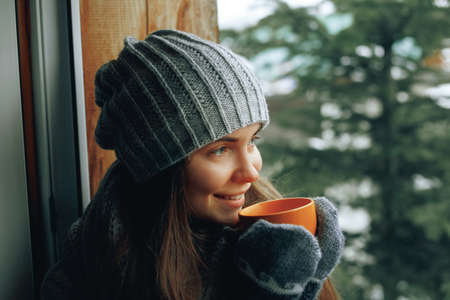 Beautiful woman holding and drinking a cup of coffee or cocoa in gloves sitting home by the window. Blurred winter snow tree background. Morning, coziness, winter and people conceptの写真素材