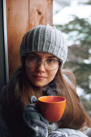 Beautiful woman holding and drinking a cup of coffee or cocoa in gloves sitting home by the window. Blurred winter snow tree background. Morning, coziness, winter and people conceptの写真素材