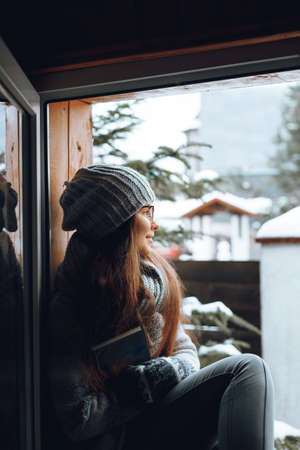 Beautiful woman in a sweater sitting home by the window. Blurred winter snow tree background. Morning, coziness, winter and people conceptの写真素材