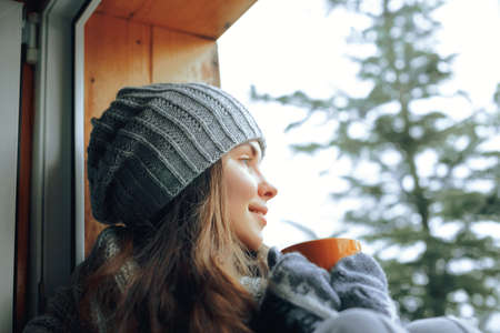 Beautiful woman holding and drinking a cup of coffee or cocoa in gloves sitting home by the window. Blurred winter snow tree background. Morning, coziness, winter and people conceptの写真素材