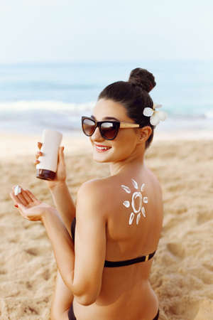 Woman Applying Sun Cream on Tanned Shoulder In Form Of The Sun. Sun Protection Sun Cream. Skin and Body Care. Girl Using Sunscreen to Skin. Female Holding Suntan Lotion and Moisturizing Sunblock.の写真素材