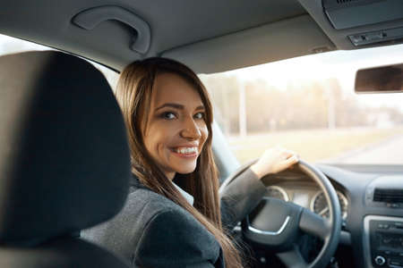 Driver turns to passenger seat and smiles at camera. Happy woman taxi driver sitting in car holding to steering wheelの写真素材