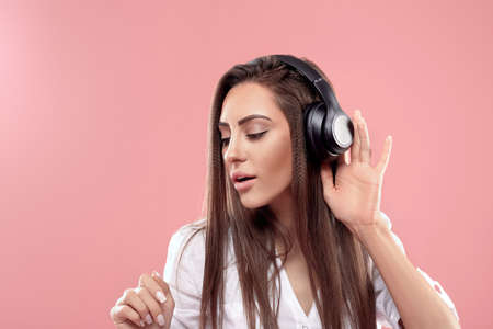 Beautiful young woman in wireless headphones listening to music on pink background. Girl uses...