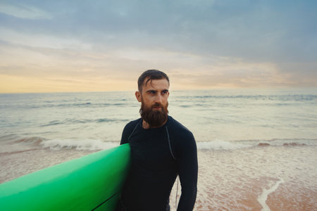 Young bearded man with surfboard standing near a beach. Man with surfing board outdoors on a summer day.の写真素材
