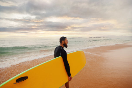 Young handsome male surfer with a surfboard is enjoying a view while walking a sandy beach at sea....