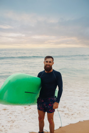 Young handsome male surfer with a surfboard is enjoying a view while walking a sandy beach at sea....