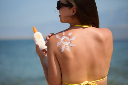 Woman Applying Sun Cream on Tanned Shoulder In Form Of The Sun. Sun Protection. Skin Care. Girl Using Sunscreen to Skin. Portrait Of Female Holding Suntan Lotion or Moisturizing Sunblockの写真素材