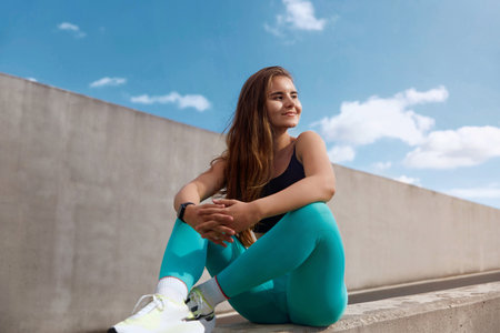 Athletic woman with long hair dressed in workout attire sits on a concrete barrier, enjoying the...