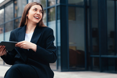 Smiling woman in a black suit holds a tablet while seated outdoors in a contemporary urban environment, expressing joy and engagement with technologyの写真素材