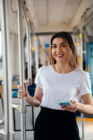 Young woman smiles while holding a smartphone in a public transport environment, enjoying her daily commute and embracing urban lifestyleの写真素材