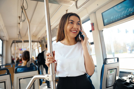 Young woman smiling and talking on her smartphone in a public transport environment with other passengers, reflecting an urban lifestyleの写真素材