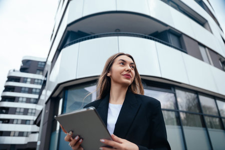 Confident businesswoman holding a tablet stands outside a modern building, highlighting sleek architecture and an urban lifestyleの写真素材