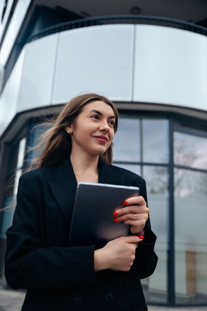 Confident young woman holding a tablet stands outside a modern building, reflecting a professional style and contemporary urban lifestyleの写真素材