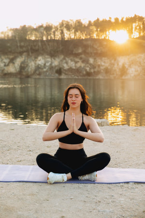 Practicing yoga on a mat by the lakeshore, a woman finds peace during sunset with warm sunlight filtering through the trees, creating a calm atmosphere.の写真素材