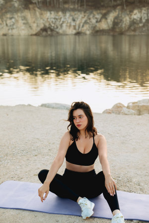 A woman is seated in a yoga pose on a mat by the lake, enjoying the serene morning view as the sun rises behind the rocky cliffs.の写真素材