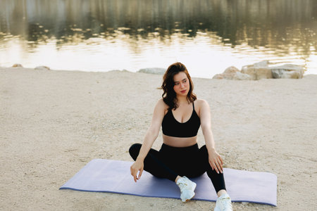 A woman sits on a yoga mat by a peaceful lake, practicing mindfulness and relaxation after a workout in the late afternoon sun.の写真素材