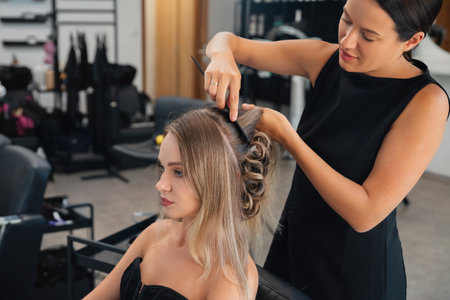 A stylist is carefully arranging a client's hair into a beautifully styled updo in a contemporary salon setting. Sunlight fills the space, enhancing the atmosphere.の写真素材
