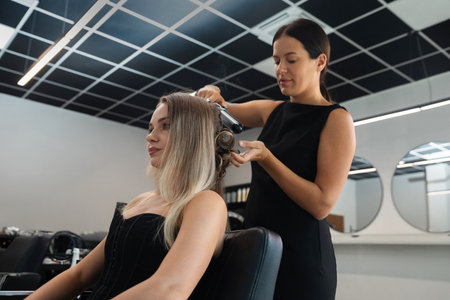 A stylist works on a client's hair, using a curling tool to create soft curls at a contemporary salon. Natural light shines through the windows.の写真素材