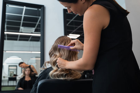 A stylist skillfully combs through a client's wavy hair in a contemporary salon. Two additional clients relax in the background while other stylists work.の写真素材
