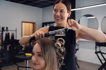 A hairstylist is curling a client's long hair with a curling iron in a sleek salon. The atmosphere is bright and contemporary, ideal for hair transformations.の写真素材