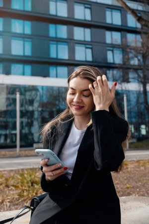 A woman with long hair sits outside, checking her phone and smiling. She wears a black blazer and enjoys a sunny day in front of a contemporary glass building.の写真素材