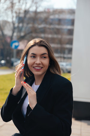 Young woman with long hair smiles while engaged in a phone conversation outside. She appears joyful, wearing casual attire in an urban setting with blurred cars in the background.の写真素材