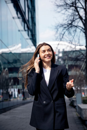 A young woman dressed in a black suit smiles brightly while speaking on her phone, walking along a city sidewalk surrounded by contemporary buildings and trees.の写真素材