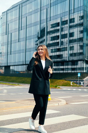 A woman dressed in a stylish outfit walks confidently across a crosswalk, talking on her phone and smiling. Modern glass buildings rise in the background.の写真素材