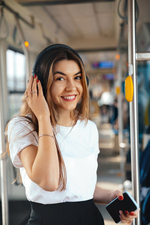 A young woman with long hair smiles while listening to music on headphones inside a city bus. She holds a smartphone and enjoys her journey amidst fellow commuters.の写真素材