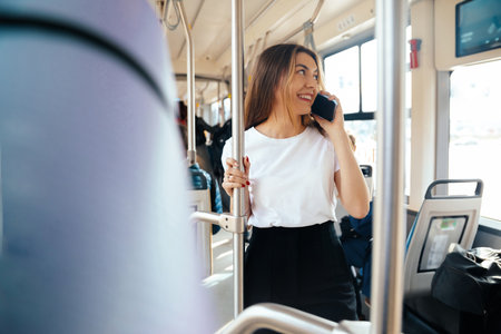 Young woman smiling and talking on her smartphone while riding public transport, embracing her urban...