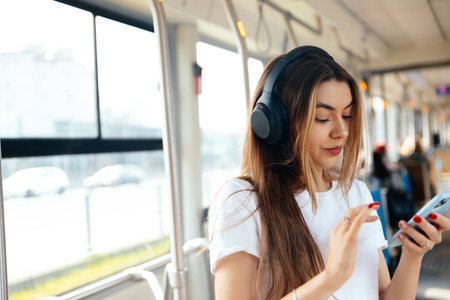Young woman listens to music through headphones while using her smartphone in a public transport environment, showcasing a modern urban lifestyleの写真素材